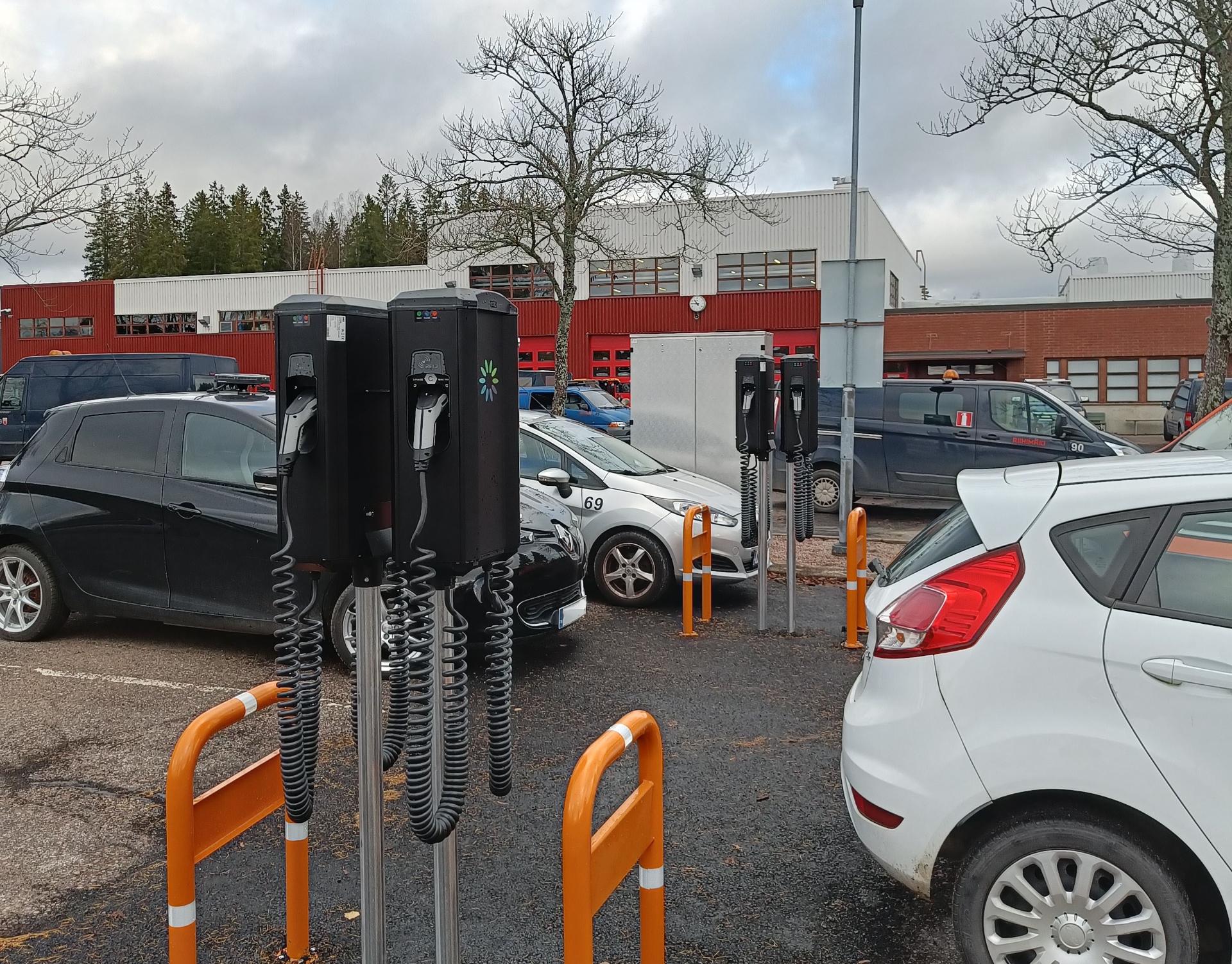 Sähköautojen latausasemia pysäköintialueella, taustalla autoja ja punavalkoinen teollisuusrakennus