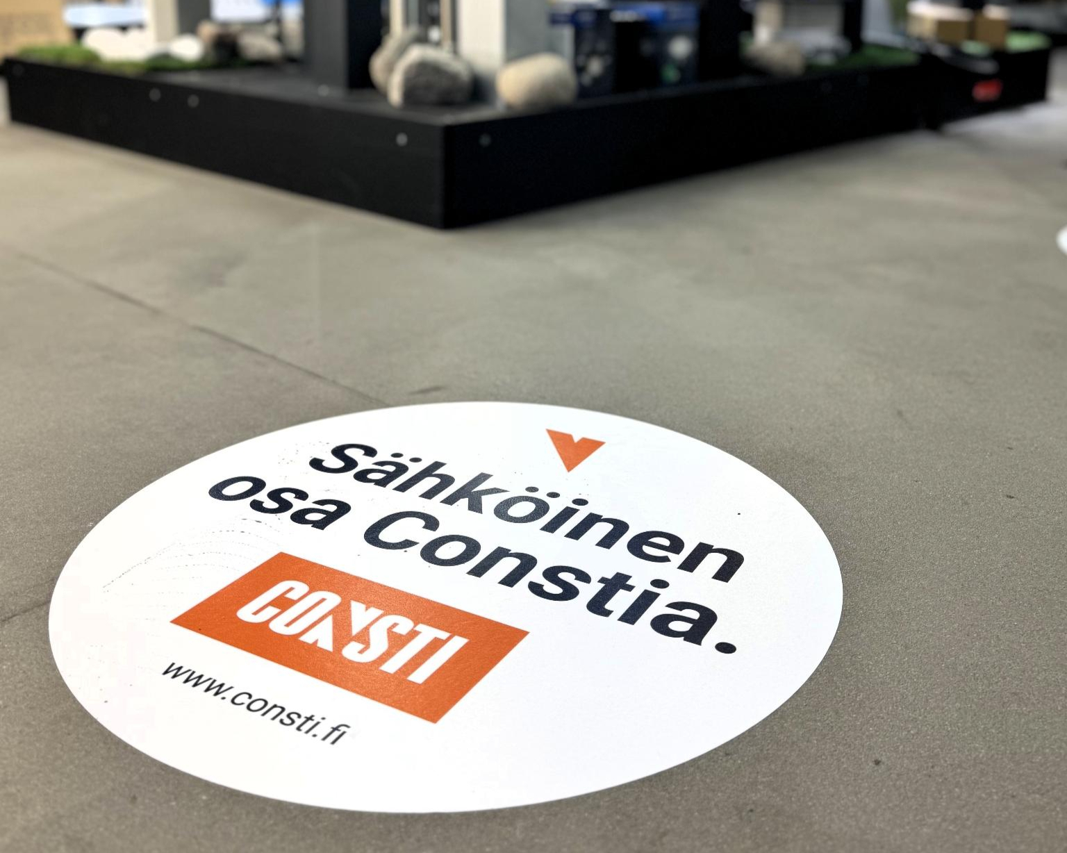 Pyöreä lattiatarra, jossa teksti Sähköinen osa Constia ja Consti-logo, taustalla tuote-esittelytasoja